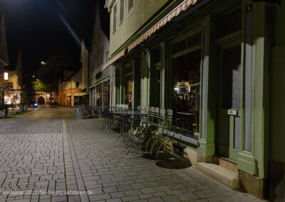 Straßenzene aus Reutlingen bei Nacht: Gepflasterte Straße, rechts Schaufenster von zwei Cafés mit hochgeklappten Stühen, im Hintergrund das beleuchtete Tor des Stadtmuseums