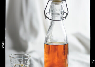 Glasflasche mit Schnappverschluss, etwa zur Hälfte mit rostroter, transparenter Flüssigkeit gefüllt. Daneben ein Schnapsglas, ebenfalls halb voll mit dieser Flüssigkeit.