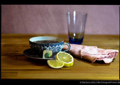 Eine Tasse mit blauem chinesischen Muster steht auf einem hölzernen Tisch. Sie ist mit Tee gefüllt, der Teebeutel hängt vorne. An der Untertasse lehnen zwei Zitronenscheiben, rechts daneben liegt eine rosafarbene Serviette. Im Hintergrund steht ein blaues Glas vor einer rosafarbenen Wand.