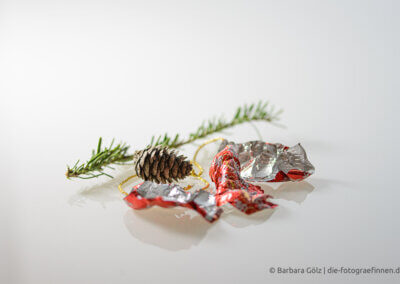 Ein einzelner Tannenzweig, ein kleiner Zapfen mit einer goldenen Schnur und das rot-silberne Einwickelpapier von Weihnachtsscholokalde liegen auf spiegelndem weißen Hintergrund