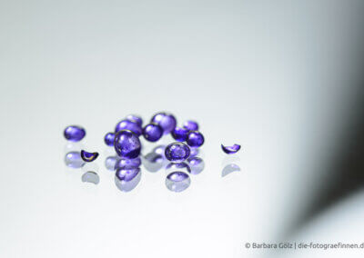 Transparente, lila gefärbte Perlen, einige davon sind zerbrochen. Sie spiegeln sich im weißen Untergrund