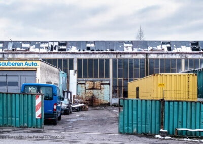 Straßenbild eines vergammelten Bauhofes mit zersprungenem Glasdach, einer rostigen Türe, einem grünen Metallzaun, hinter dem ein gelber Container und ein blaues Auto stehen. Auf dem Dach liegen Schneereste