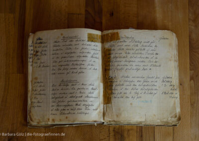 Ein sehr altes Buch mit handschriftlich eingetragenen Rezepten liegt auf einer Holzplatte. Die Seiten sind vergilbt und fleckig, in der Mitte ist es mit Klebeband geflickt.