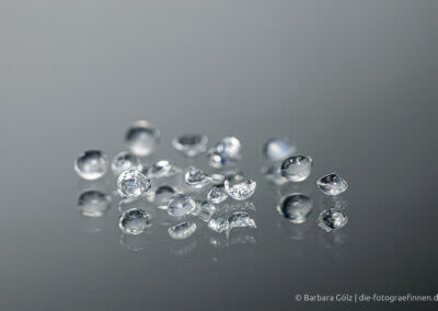 Transparente Perlen, einige davon sind zerbrochen. Sie spiegeln sich im grauen Untergrund